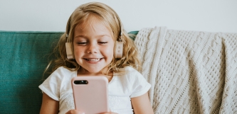 Ein Mädchen sitzt mit Smartphone und Kopfhörern fröhlich strahlend auf dem Sofa.